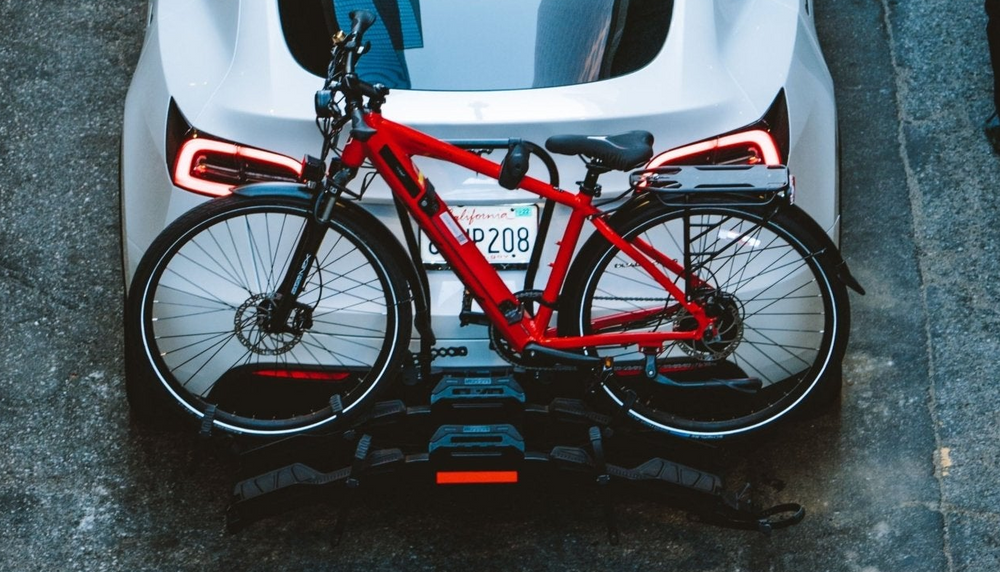 A red bicycle mounted on a black rack attached to the rear of a white car, photographed from above on a paved street. Part of a person is visible standing at the upper right.