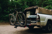 White truck with a bike rack and a tent on the back, parked in a forest.